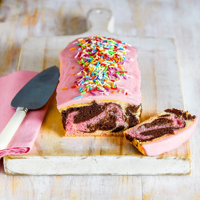 gluten-free low FODMAP marble loaf cake with pink icing and sprinkles on a wooden board with a slice to the side.