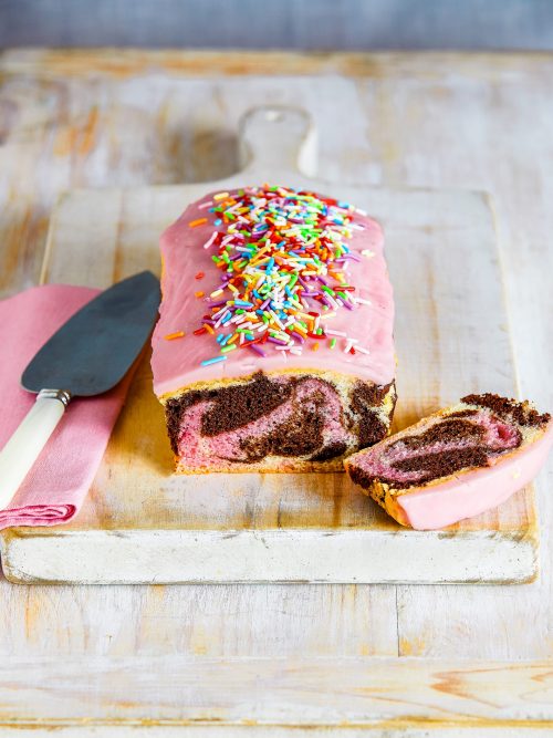 gluten-free low FODMAP marble loaf cake with pink icing and sprinkles on a wooden board with a slice to the side.