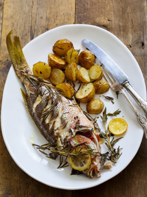 whole roast snapper on a white plate with roasted potatoes and herbs.