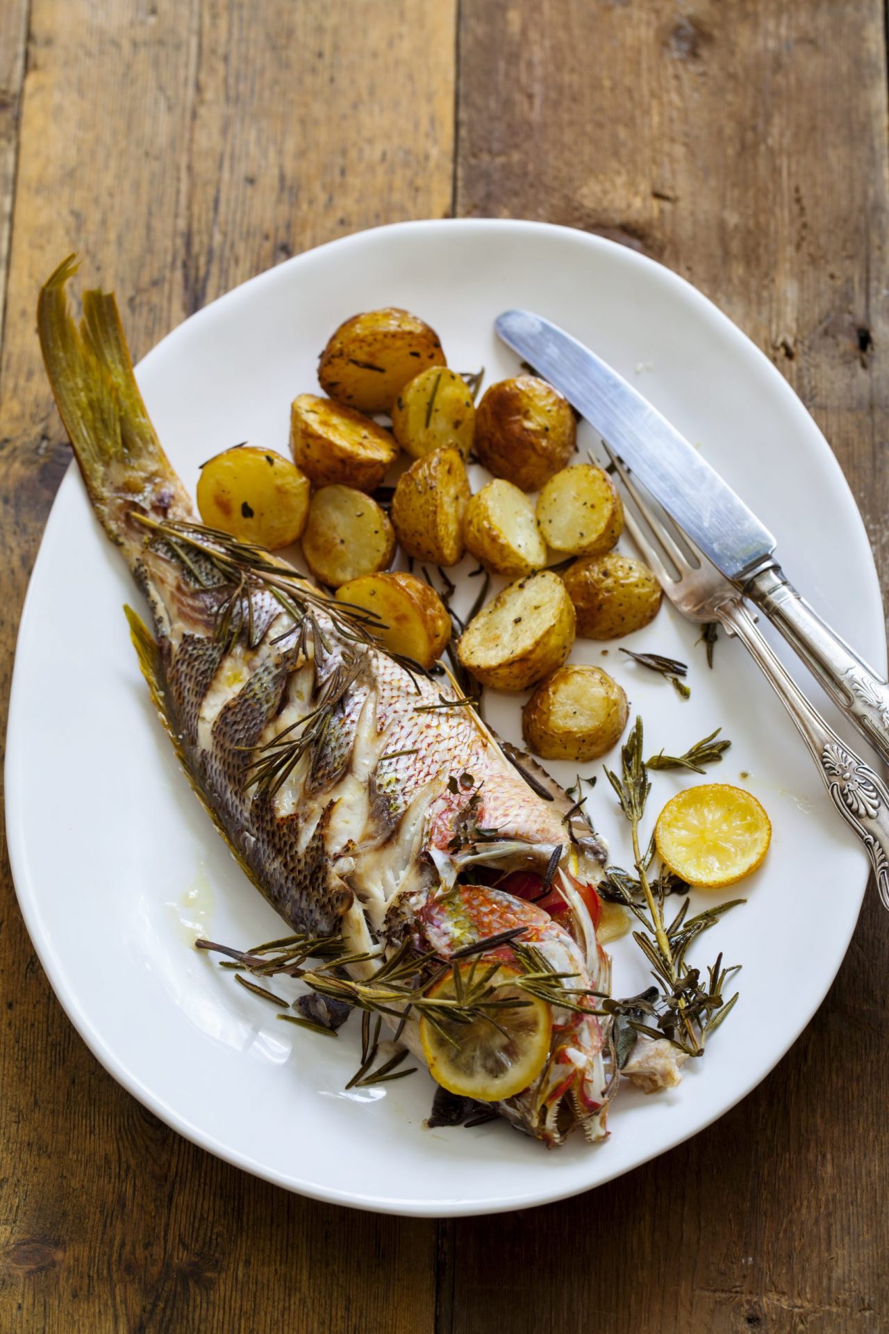whole roast snapper on a white plate with roasted potatoes and herbs.