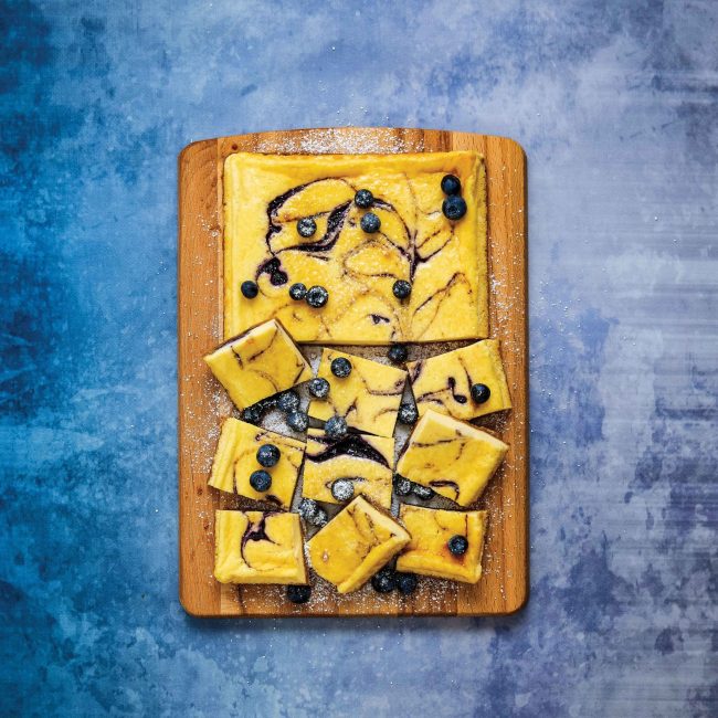 Low FODMAP Blueberry cheescake slice on a wooden board, partially sliced into squares. On a blue background.