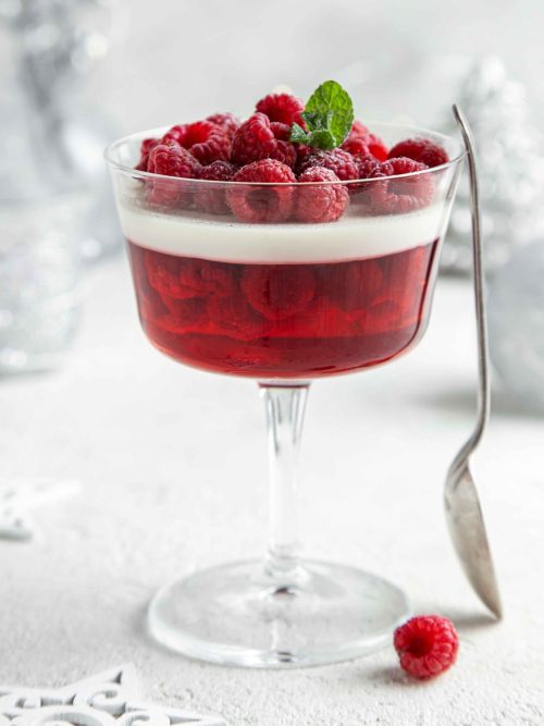 Low FODMAP Berry Coconut Panna Cotta served in an large stemmed glass. Topped with fresh raspberries and mint leaves. Spoon resting on the side. In the background are white and silver Christmas decorations.