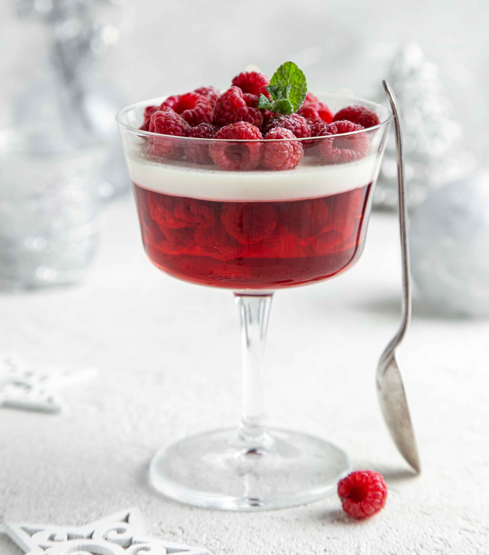 Low FODMAP Berry Coconut Panna Cotta served in an large stemmed glass. Topped with fresh raspberries and mint leaves. Spoon resting on the side. In the background are white and silver Christmas decorations.