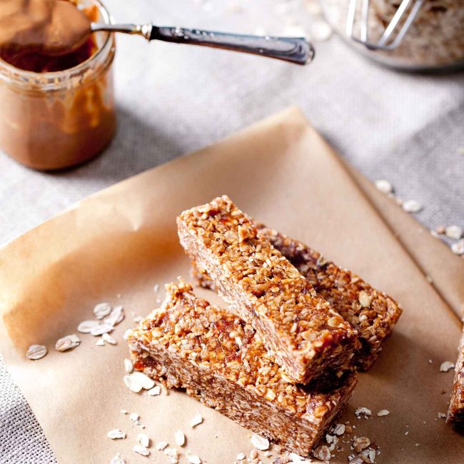Three low FODMAP no-bake peanut butter oat bars stacked on baking paper with jars of peanut butter and oats in the background.