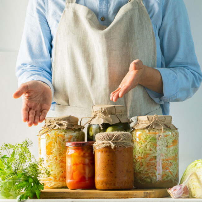 Picture of a cook in a light blue shirt and white apron pointing to jars of assorted homemade fermented vegetables and pickles