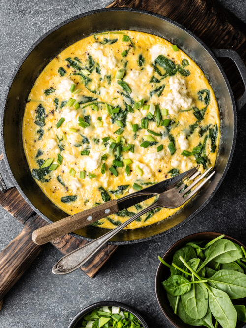 Low FODMAP Spinach & Feta Omelette in a frying pan ready to serve. Shot top down with a bowl of spinach at the side and a knife and fork ready to serve.