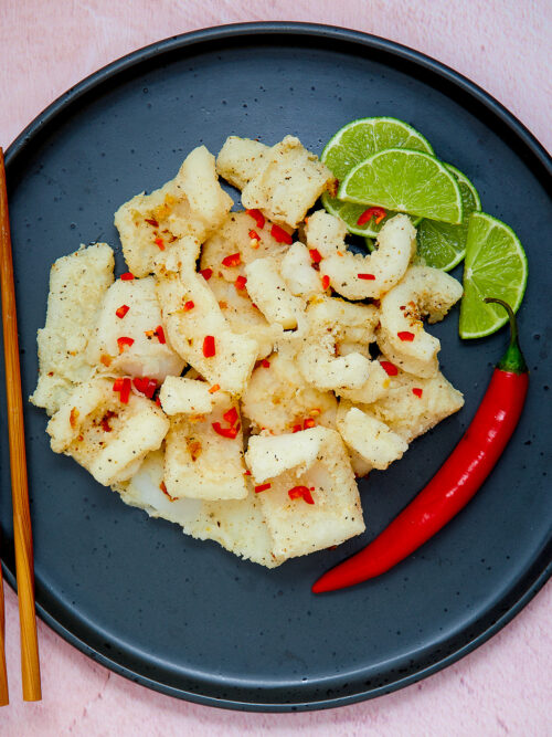 A black plate is piled with pieces of Monash endorsed Salt and Pepper Squid. Pieces of lime and a fresh chilli sit on the side.