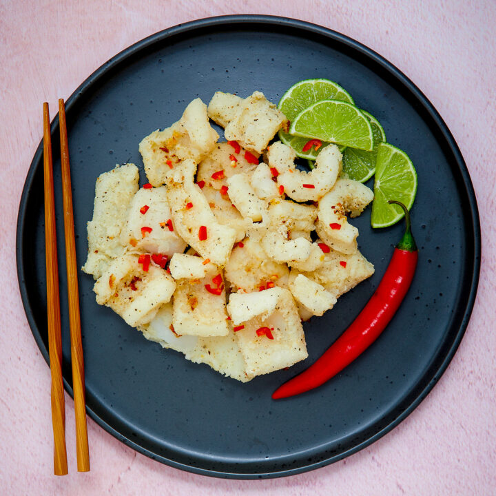 A black plate is piled with pieces of Monash endorsed Salt and Pepper Squid. Pieces of lime and a fresh chilli sit on the side.