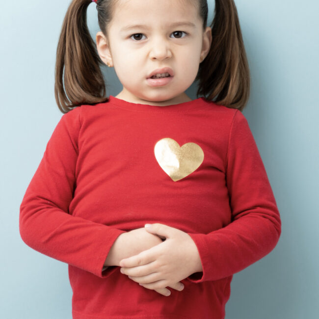 Young girl in long-sleeved red top with a gold heart. Has stomach ache. Hands are on tummy.