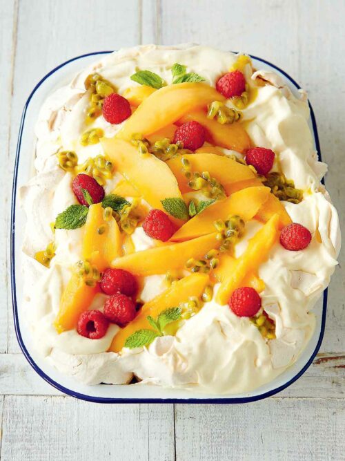 Low FODMAP Slab Pavlova, also known as Tray-Bake Pavlova in an enamel baking dish with blue edge. Toppwed with fresh cream, mango slices, fresh raspberries, passionfruit pulp and mint leaves.