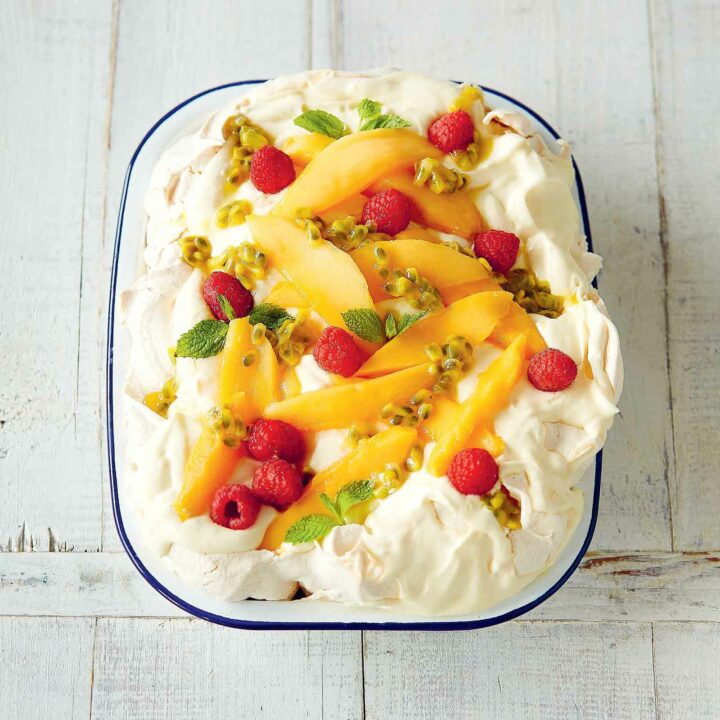 Low FODMAP Slab Pavlova, also known as Tray-Bake Pavlova in an enamel baking dish with blue edge. Toppwed with fresh cream, mango slices, fresh raspberries, passionfruit pulp and mint leaves.