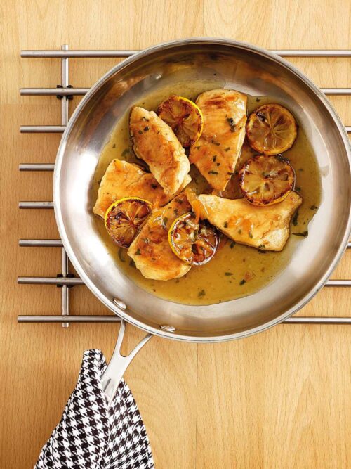 A frying pan with cooked chicken fillets garnished with caramelized orange slices and coated in a golden orange sauce. The pan rests on a metallic trivet placed on a wooden surface, with a black and white checkered cloth draped over the handle.