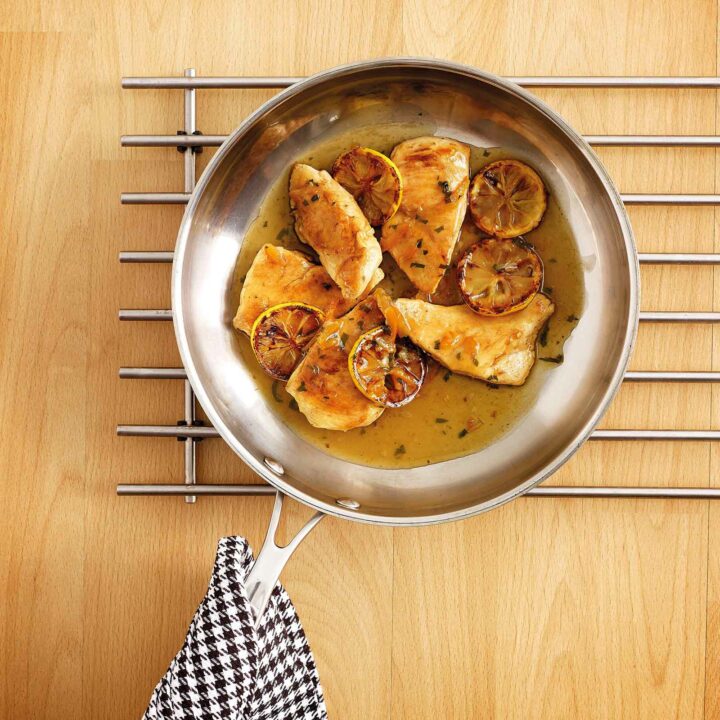 A frying pan with cooked chicken fillets garnished with caramelized orange slices and coated in a golden orange sauce. The pan rests on a metallic trivet placed on a wooden surface, with a black and white checkered cloth draped over the handle.