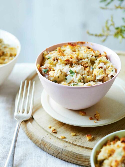 A serving of Low FODMAP Chicken Mornay in a small pink bowl with a crispy golden breadcrumb topping. The bowl is placed on a wooden board with a fork beside it, showcasing the creamy and comforting dish.