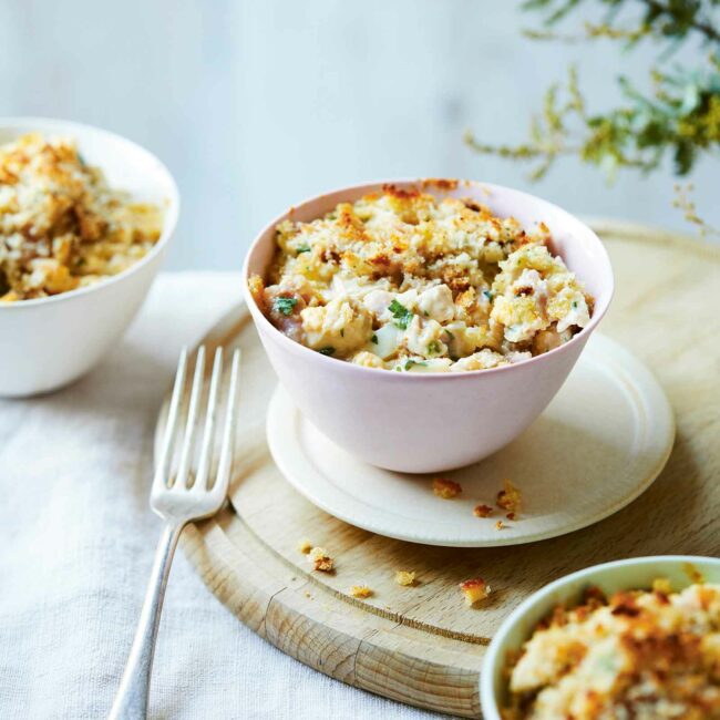 A serving of Low FODMAP Chicken Mornay in a small pink bowl with a crispy golden breadcrumb topping. The bowl is placed on a wooden board with a fork beside it, showcasing the creamy and comforting dish.